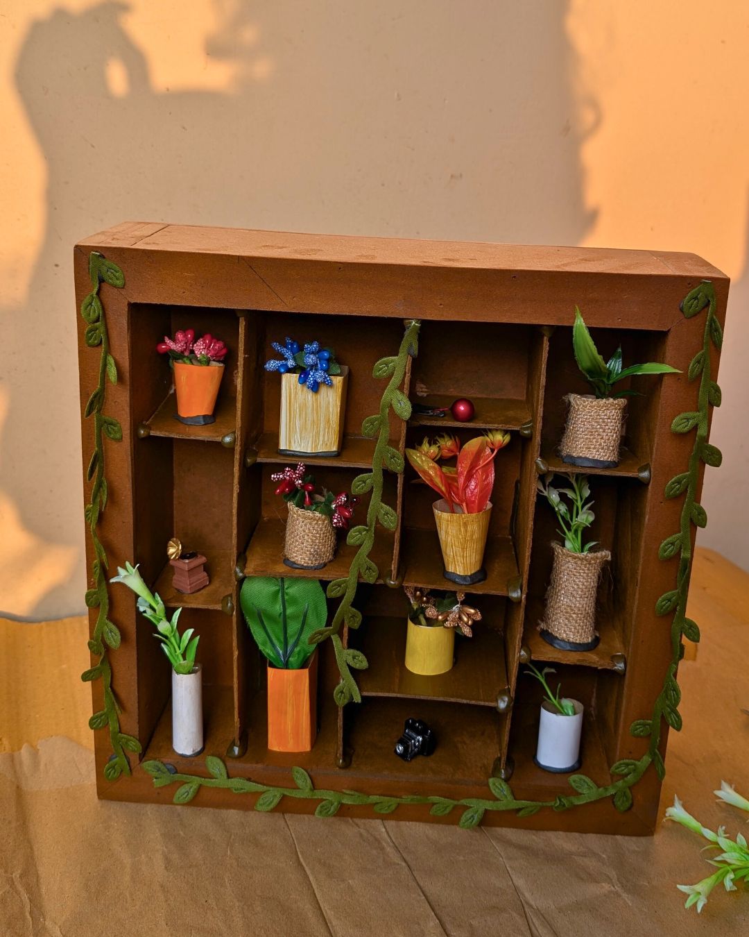 Miniature wooden frame titled "A House of Vases" featuring handcrafted mini vases and planters arranged on brown shelves, decorated with vine leaves.