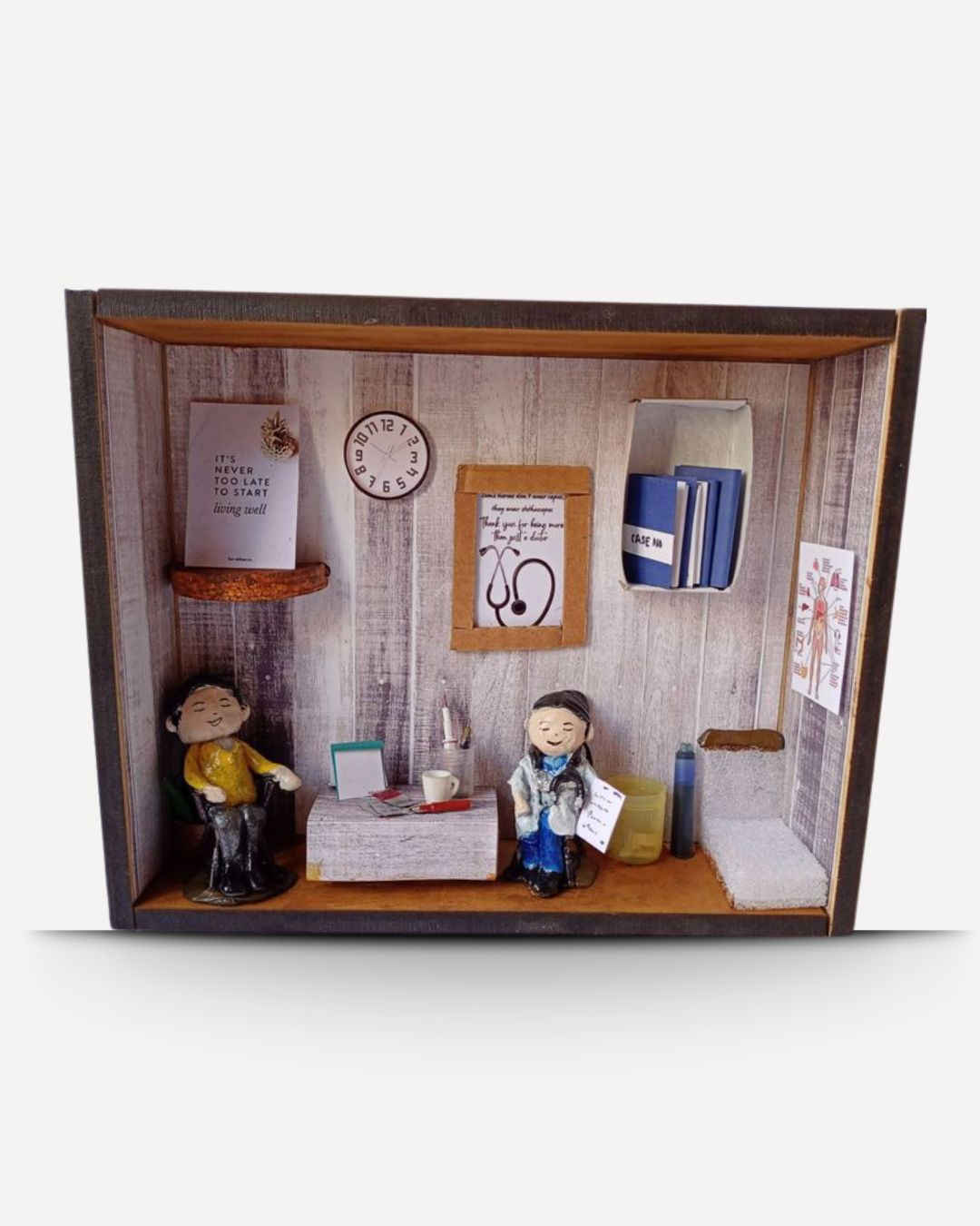 A miniature diorama model of a doctor's office, featuring small figurines of a doctor and patient, a desk with a stethoscope, medical files, and wall decorations, all enclosed in a wooden frame.