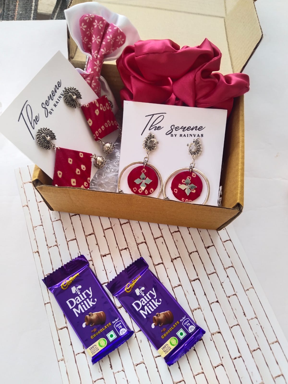 Brown box with multiple earrings, chocolates, dry fruits, hair scrunchies on white backdrop