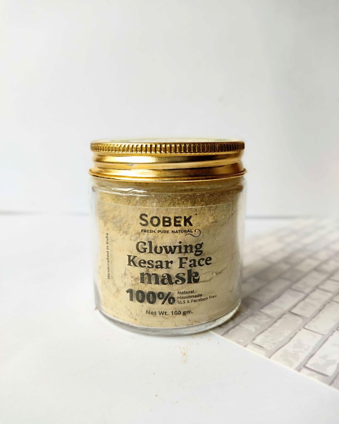 Glass jar with sobek brown kesar face mask powder on a white backdrop