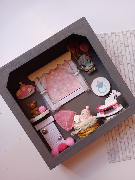 Wooden grey shadow box frame with miniature baby girl props in pink and white