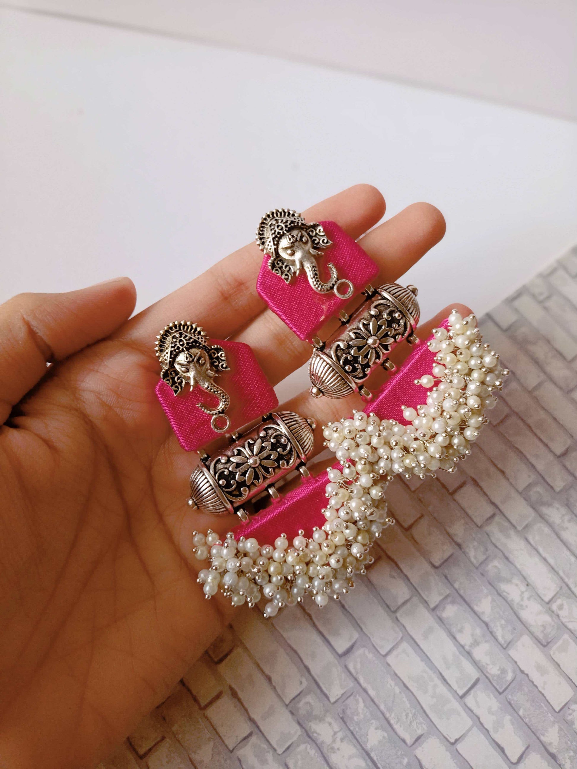 Palm holding pink earrings with ganpati face charm and silver white beads at the bottom on white grey backdrop