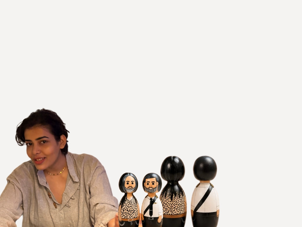 A woman with dark hair wearing a light beige button-up shirt is seated at a table, looking toward the camera. In front of her are four hand-painted wooden nesting dolls, including two with black hair and leopard-print clothing and two with white clothing.