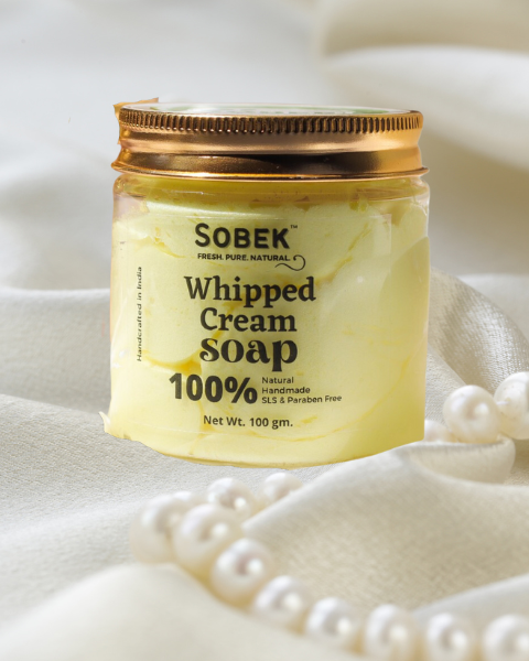 Glass jar with sobek yellow whipped cream soap, pearl necklace on one side on white cloth
