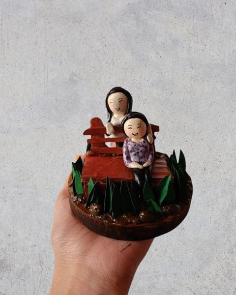 Hand holding a clay miniature sculpture of two happy girls sitting on bench
