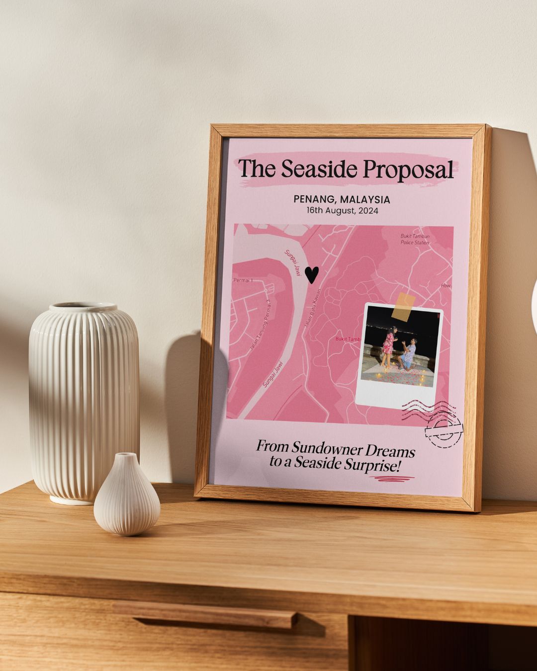 Framed map of Penang with a couple on a beach, placed on a wooden surface with decorative vases.