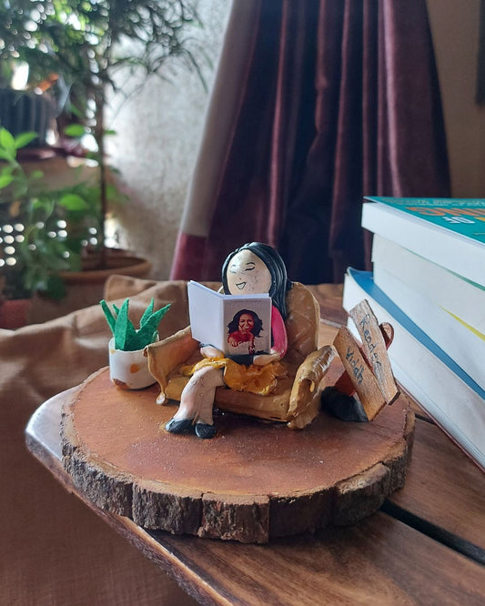 A small decorative figurine of a seated woman reading a book, displayed on a round wood slice tray. The scene includes books, a small potted plant, and curtains in the background.