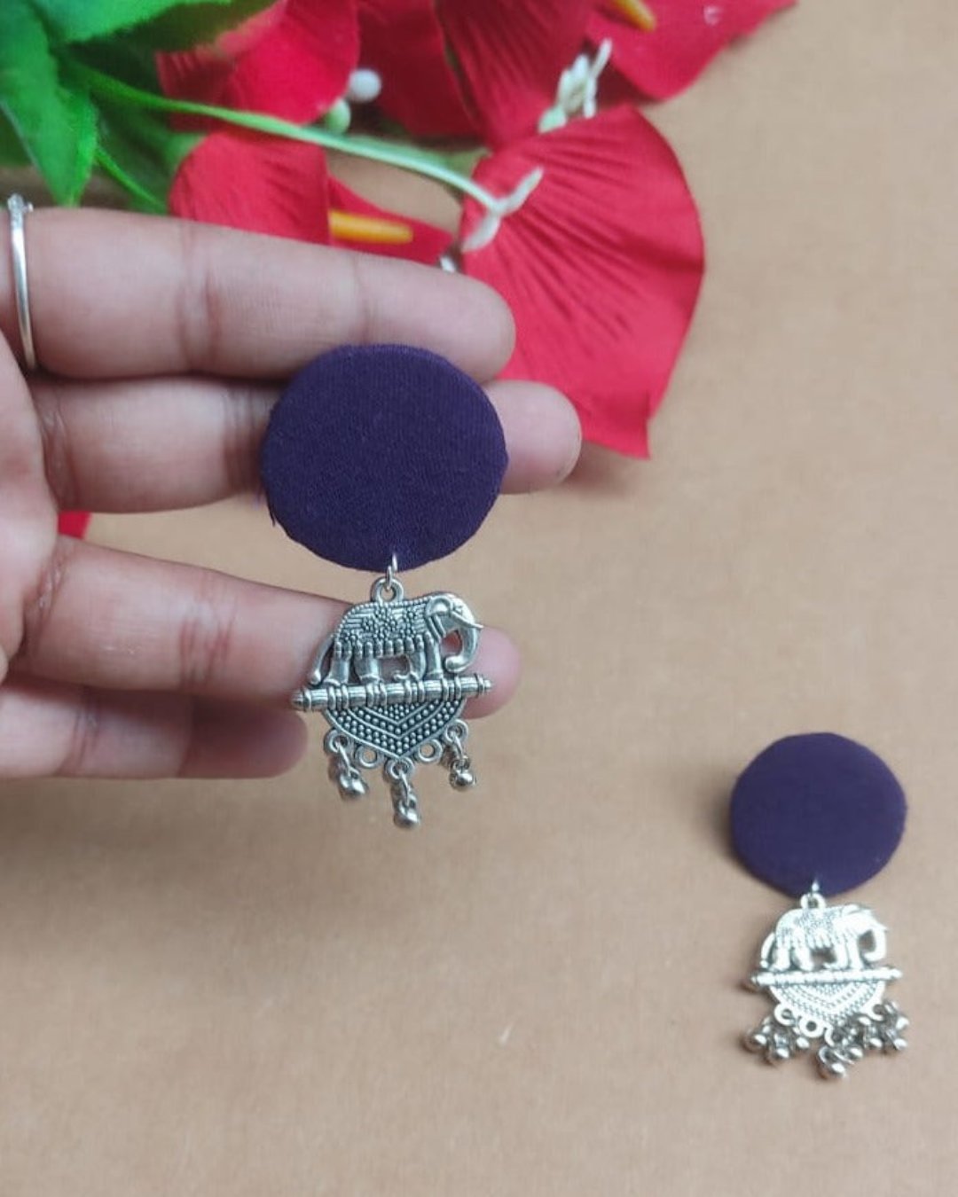 Fingers holding Blue round earrings with silver charm at bottom on brown backdrop