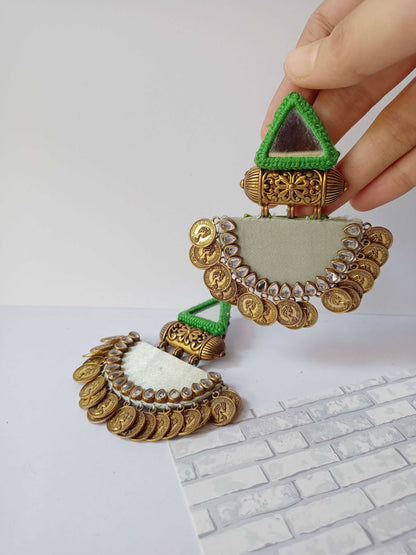 Hand holding dark and light green jhumka earrings with golden coins and kundan on white grey backdrop