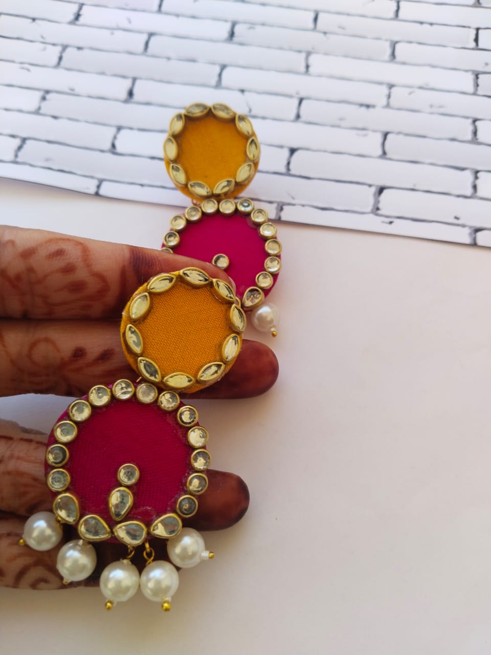 Finders holding Pink and yellow round earrings with kundan border and white pearls on white grey backdrop