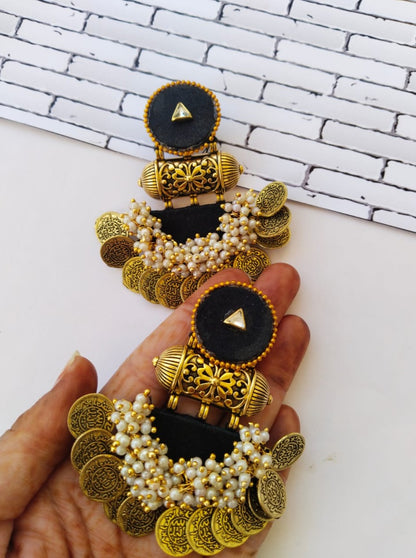 Hand holding black heavy jhumka earrings with golden tabiz and coins with white beads on white backdrop