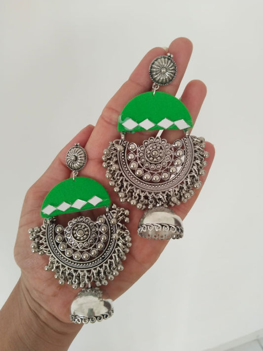 Hand holding light green heavy earrings with silver charm and ghungroos on white backdrop