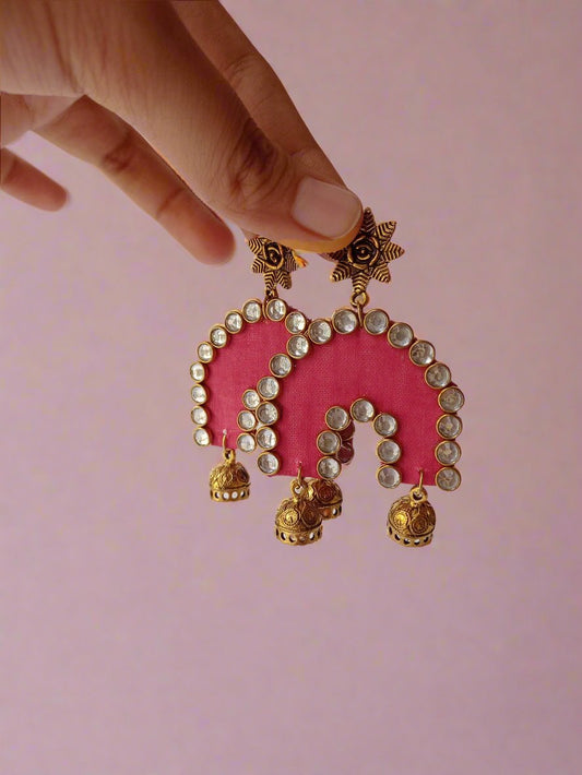 Hand holding pink semi round jhumka earrings with kundan border and golden jhumki on white grey backdrop