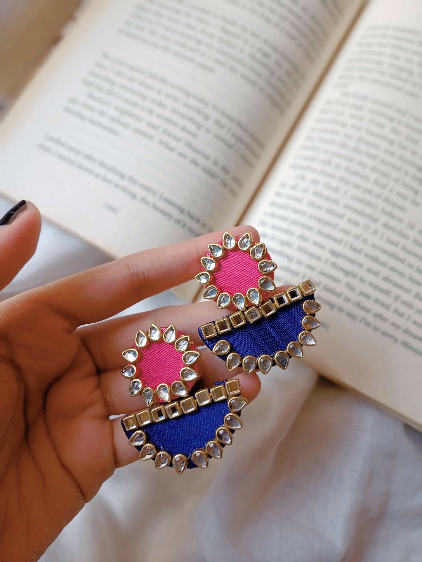 Fingers holding Pink and blue round and semi circle earrings with kundan border kept on open book backdrop