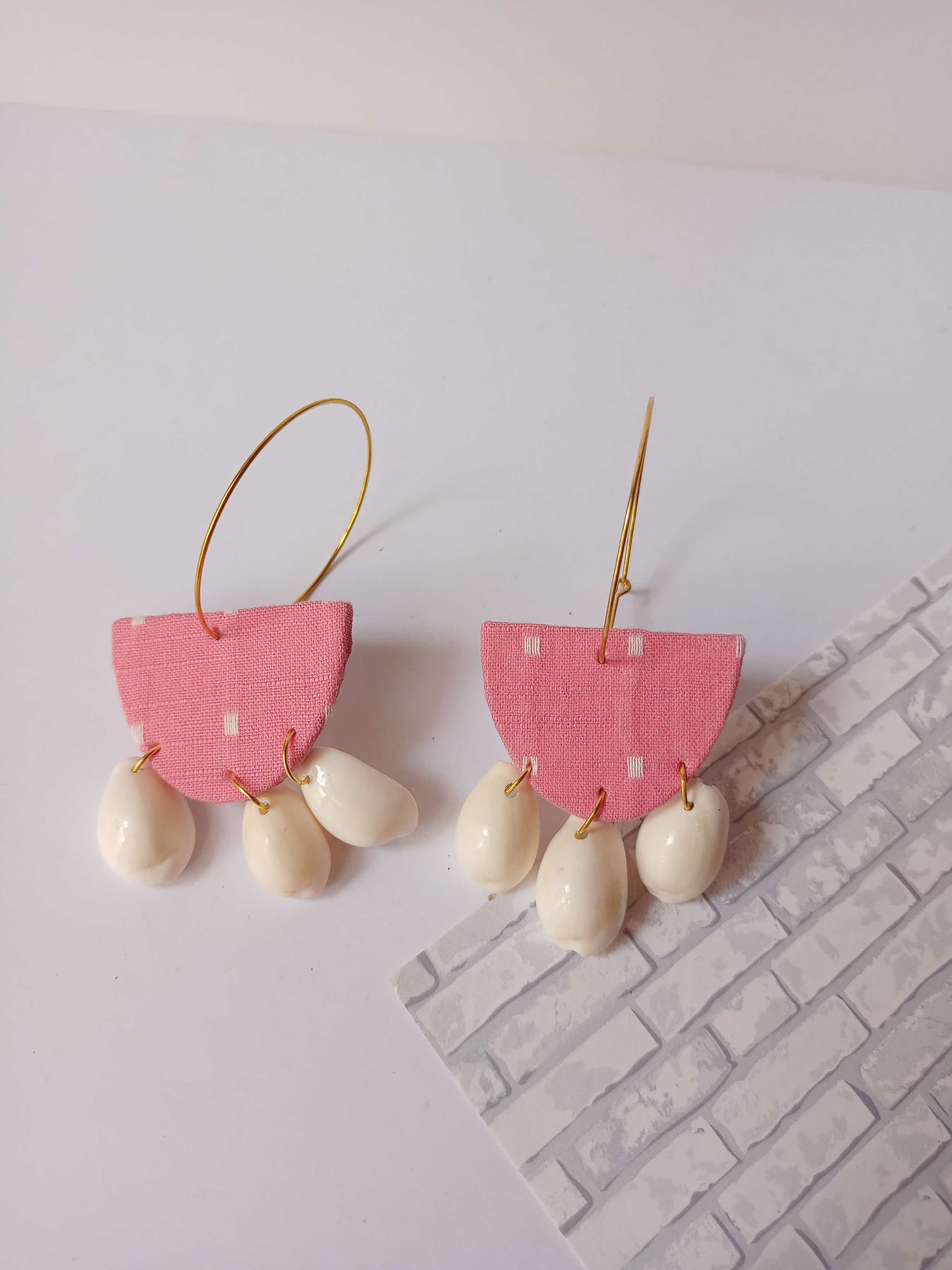 Pink semi round hoops earrings with white sea shells at the bottom on white grey backdrop