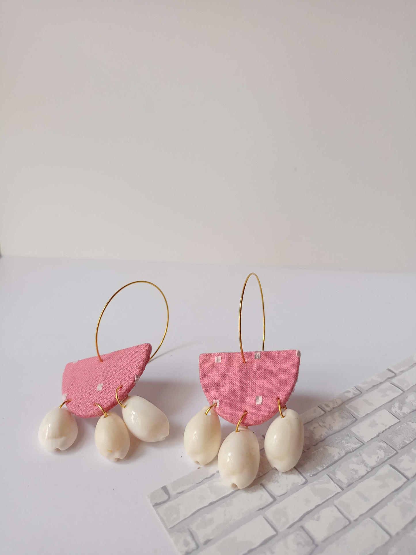 Pink semi round hoops earrings with white sea shells at the bottom on white grey backdrop