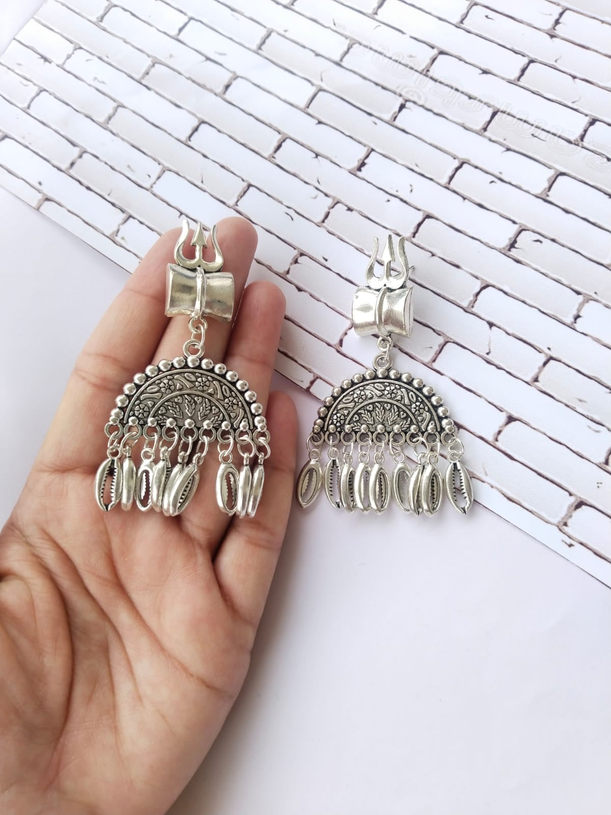 Palm holding Silver oxidized semi round jhumka earrings with silver beads and trishul charm on white backdrop